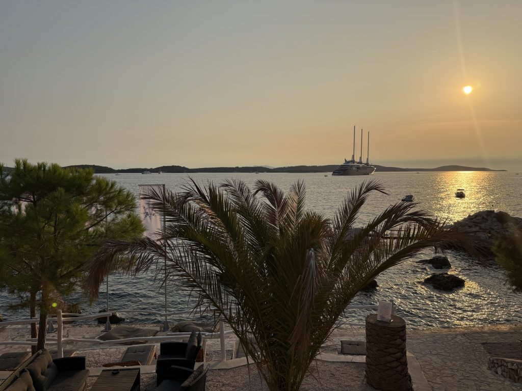 Golden sunset over the Adriatic from a seaside terrace in Hvar, with a palm tree silhouetted in the foreground, a large three-masted cruise ship anchored offshore, and low wooded islands on the horizon.