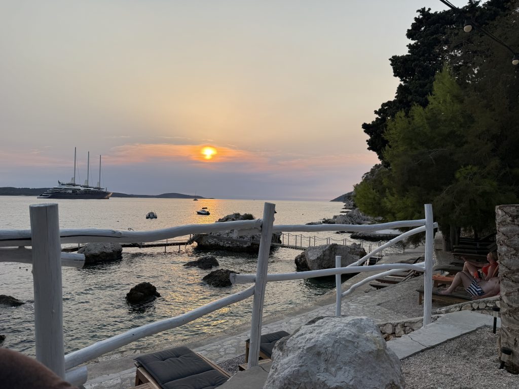 Sunset from the outdoor seaside bar at Hotel Podstine in Hvar Town, with a white driftwood railing, rocky swimming platforms, and a three-masted ship anchored offshore