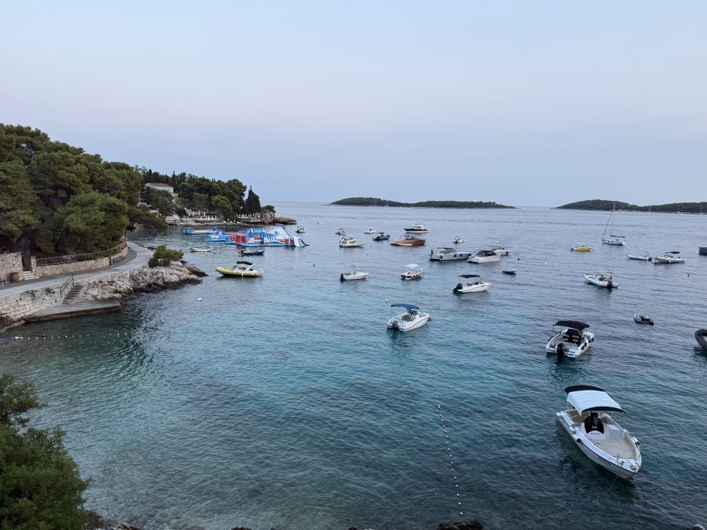 Boats anchored in a clear turquoise bay near Hvar Town at dusk, with a seaside promenade, an inflatable water park, and the Pakleni Islands on the horizon