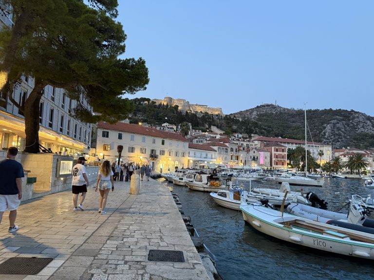 Hvar Town’s harborfront promenade at dusk, with small boats moored along the stone quay, warmly lit waterfront buildings, and the illuminated Fortica fortress on the hillside above.