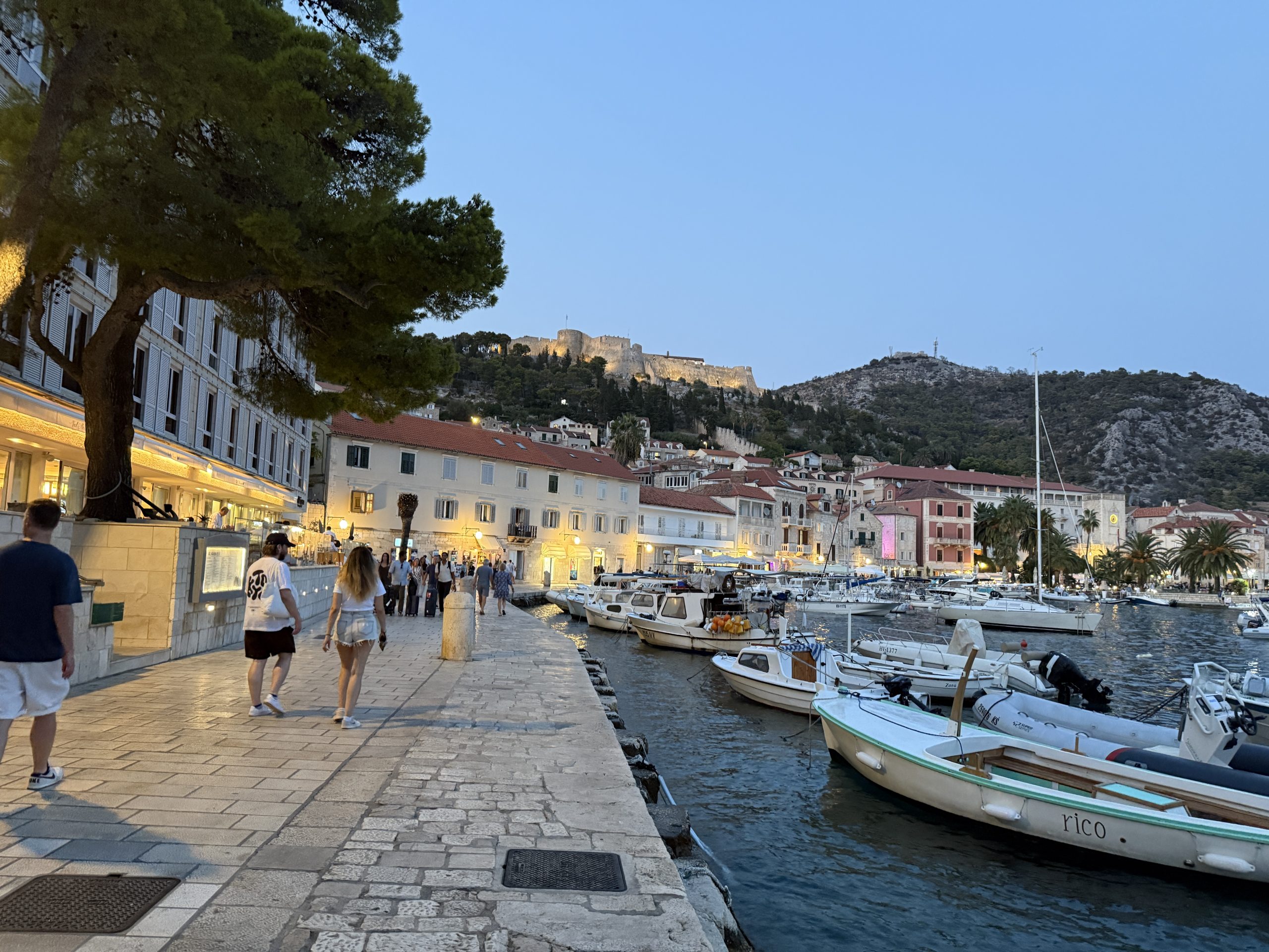 Hvar Town’s harborfront promenade at dusk, with small boats moored along the stone quay, warmly lit waterfront buildings, and the illuminated Fortica fortress on the hillside above.