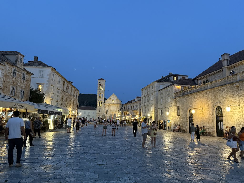 Evening crowds on the stone piazza of St. Stephen’s Square, with the cathedral’s illuminated bell tower at the far end and restaurants spilling onto the square.