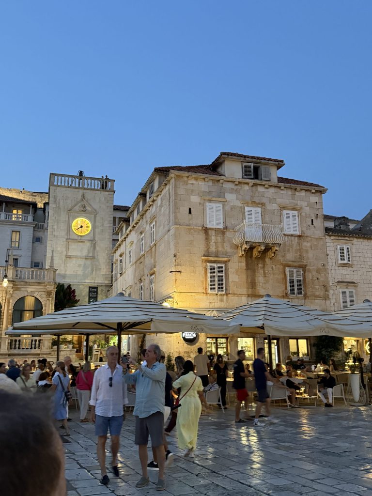 St. Stephen’s Square in Hvar Town at dusk, with the clock tower and loggia beside a historic stone building with a curved white balcony, and people strolling past café tables under large umbrellas.