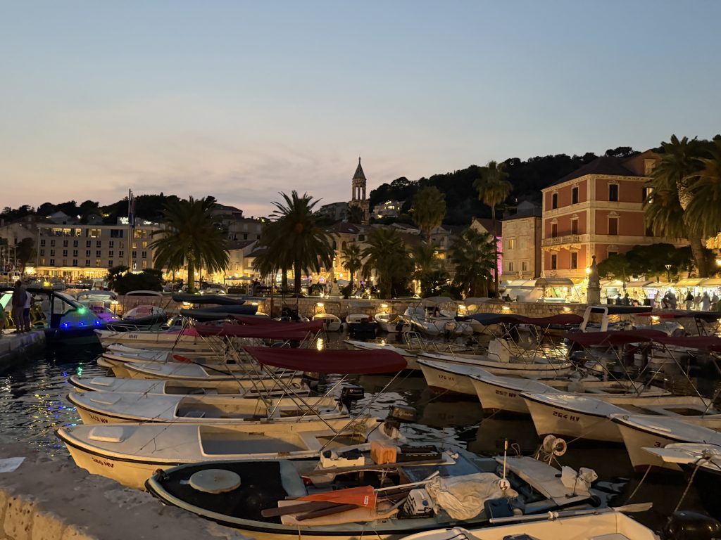 Rows of small wooden boats with maroon canopies packed into Hvar Town’s harbor at dusk, with the palm-lined waterfront, glowing shops and restaurants, and a church bell tower against a pink and blue sky.