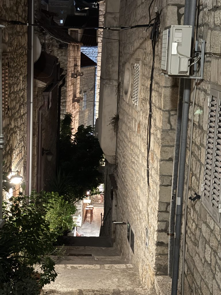 Narrow stone staircase alley winding downhill between old buildings in Hvar Town at night, lit by wall lanterns and lined with leafy plants, with café stools glowing at the bottom.