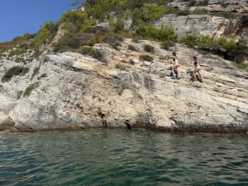 Two swimmers climbing a sloped limestone cliff above clear turquoise water at a secret swim cove on a boat tour from Hvar, Croatia.