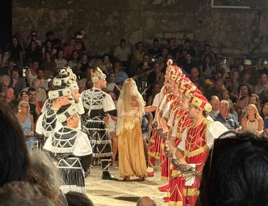 Moreška sword dance performance in Korčula, Croatia, at night. A veiled woman in flowing gold harem pants and a sheer gold veil stands between two lines of dancers. On the left, dancers in black and white costumes and ornate headdresses face her, led by a man wearing a gold crown. On the right, dancers in red and gold costumes stand in a row holding basket-hilted swords. A large audience watches from tiered seating against an old stone wall