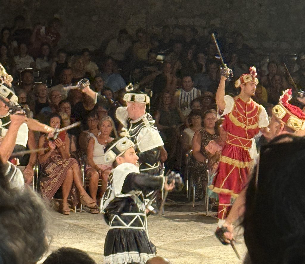 Moreška sword dancers in Korčula, Croatia, perform at night before a packed crowd. Dancers in red and gold costumes face dancers in black and white, their basket-hilted swords crossed between the two lines. Both sides wear ornate jeweled headdresses, and one dancer in the red army wears a gold crown.