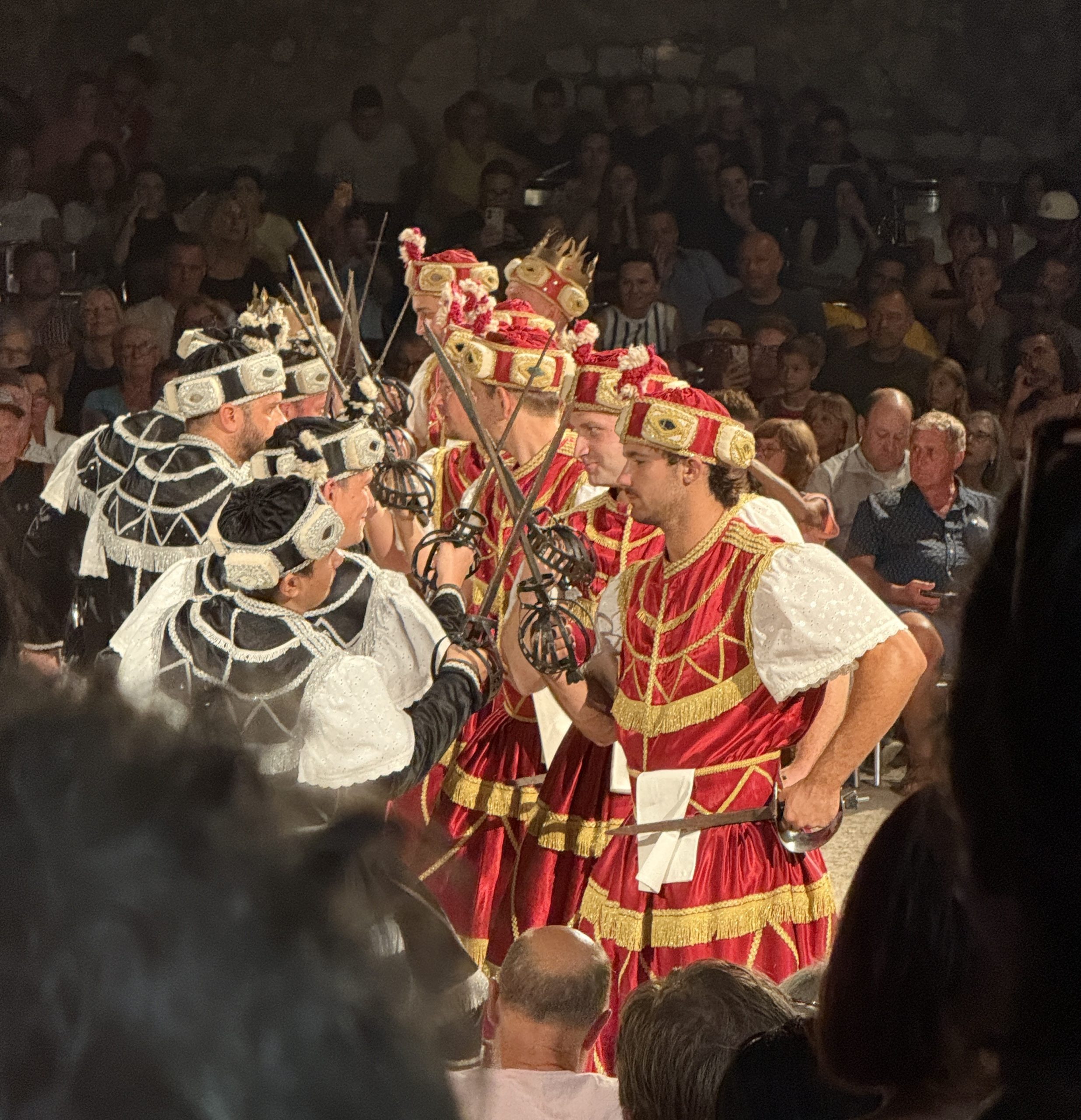 Moreška sword dancers in Korčula, Croatia, perform at night before a packed crowd. Dancers in red and gold costumes face dancers in black and white, their basket-hilted swords crossed between the two lines. Both sides wear ornate jeweled headdresses, and one dancer in the red army wears a gold crown
