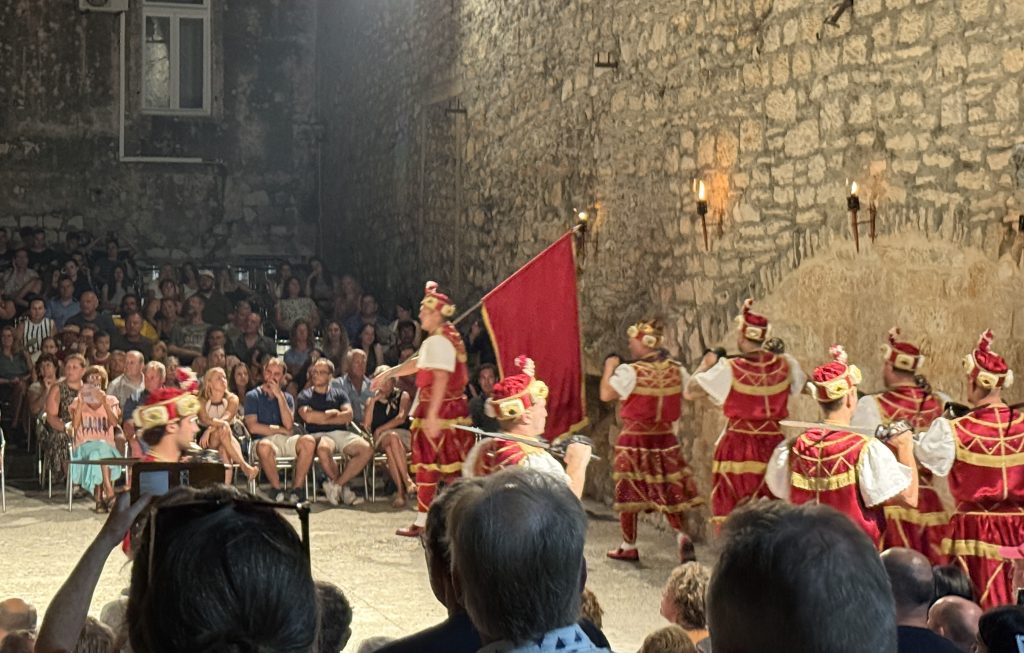 Dancers in the red army of the Moreška sword dance march in a line across a stone courtyard in Korčula, Croatia, carrying swords over their shoulders. One dancer leads with a large red flag. Behind them, lit torches hang on a tall stone wall, and audience members watch from chairs along the edge of the performance space.