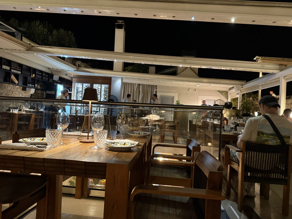 Open-air rooftop dining room at Le Specialità in Hvar Town at night, with a wooden table set with crystal glasses and hand-painted plates beside a glass railing. The railing surrounds an opening in the center of the floor that looks down to the level below. Diners sit under a white pergola open to the night sky, next to a wall of glass-fronted wine fridges and bottle shelves.