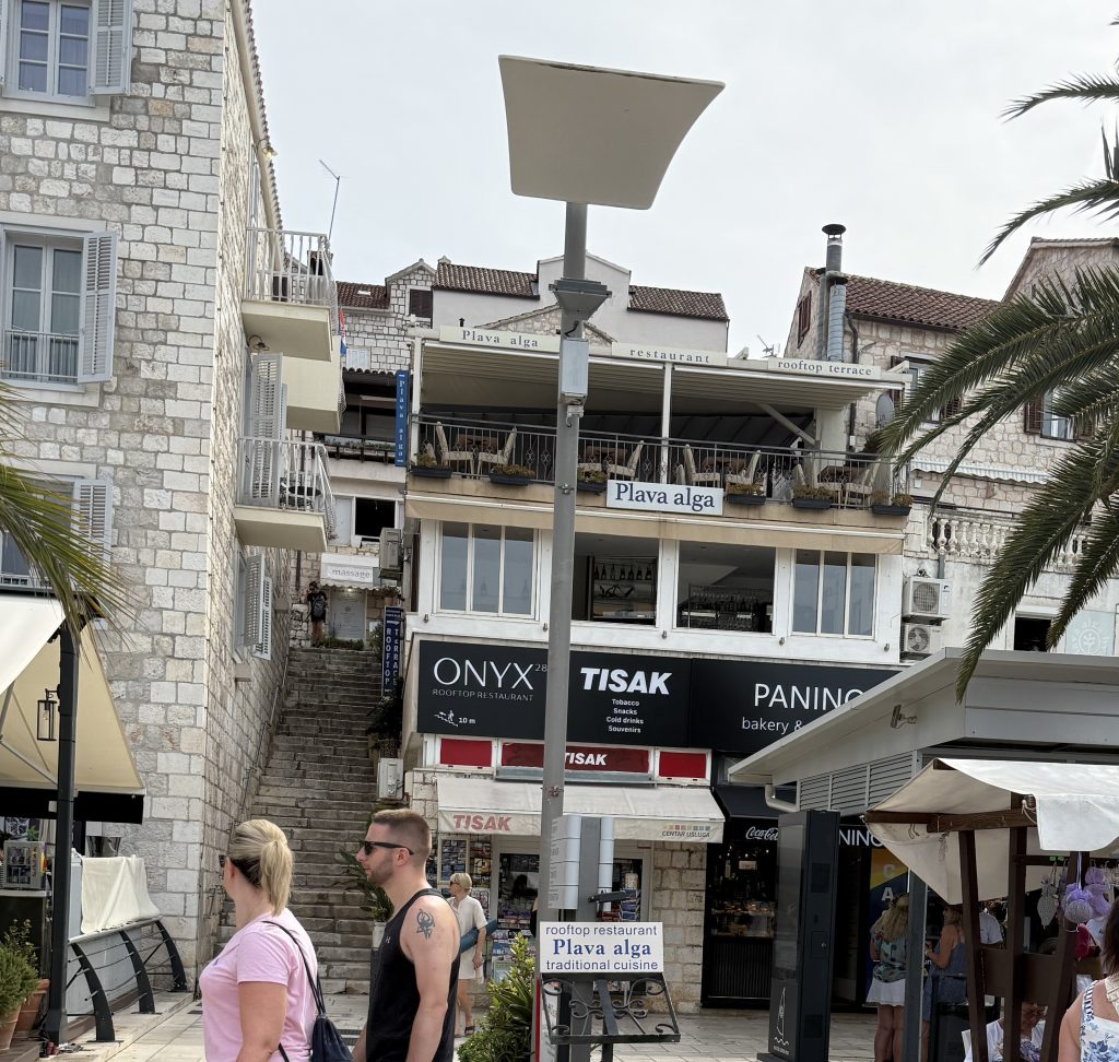 The sign for Onyx 28 Rooftop Restaurant above the shops on Hvar Town’s waterfront, with an arrow showing it’s 10 meters up the steep stone staircase beside the building. People walk past palm trees and market stalls on the promenade below.