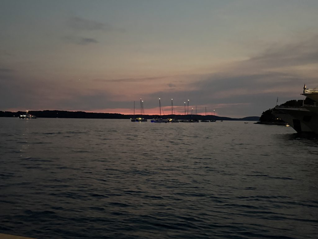 Twilight over the sea in Hvar Town, with sailboats anchored offshore, their mast lights glowing against a fading pink glow above the dark outline of the Pakleni Islands. The bow of a large yacht moored along the waterfront edges into the right side of the frame.