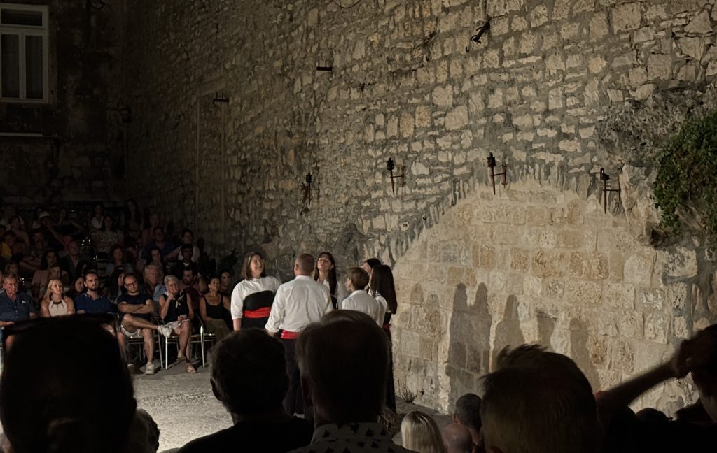 A small group of performers in white shirts, black trousers or skirts, and red sashes stand in a loose circle on the stone floor of the Moreška venue in Korčula, Croatia. Their shadows fall on the tall stone wall behind them, where unlit torches hang in a row and ivy grows near the top. Audience members watch from chairs on the left, with the heads of people in the front rows silhouetted in the foreground