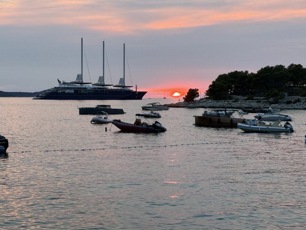 Sunset over the sea at Hvar Town, the red sun dipping into the water beside a pine-covered point. Small motorboats and dinghies float in the foreground, and a large three-masted ship is anchored offshore under a pink and lavender sky.