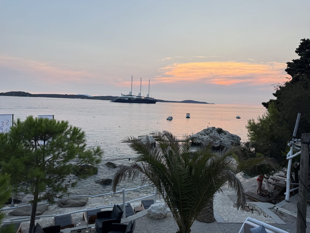 Sunset view from a seaside lounge bar in Hvar Town, looking past a palm tree and pine trees to a calm sea with a large three-masted ship anchored offshore. Low wooded islands line the horizon under a pink and orange sky, and below, a rocky shore and sunbeds sit next to a small swimming platform on a rock.