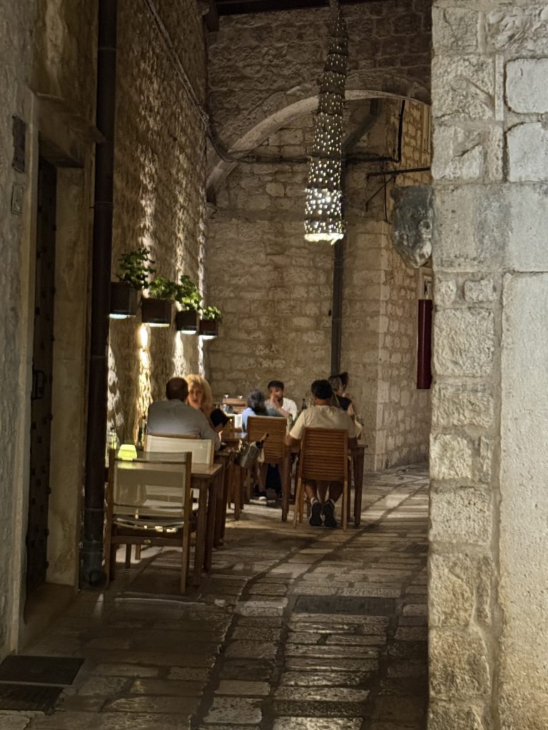 Diners at small wooden tables in a narrow stone side street in Hvar Town, lit by a hanging pierced-metal lantern and wall planters of fresh herbs, before the evening crowds arrive. A carved stone face sits on the corner of an old limestone wall beside them.