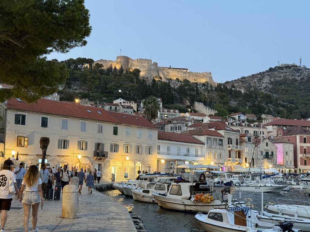 Hvar Town harbor at dusk, with people strolling the stone waterfront past small wooden fishing boats moored along the quay. Cream and pastel buildings with red-tiled roofs and glowing lanterns line the water, and the floodlit Spanish Fortress sits on the pine-covered hill above, with old town walls climbing the slope toward it.