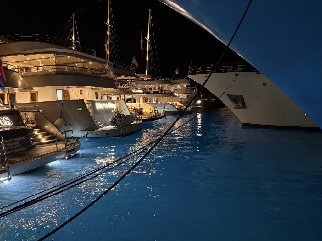 Small cruise ships and yachts flying Croatian flags, moored side by side in Hvar Town’s harbor at night, their decks glowing with warm light. Underwater lights turn the water a vivid blue, with small fish visible near the surface, while thick mooring ropes cross the foreground below the curved bow of another ship.